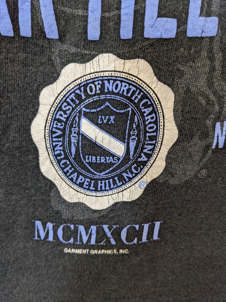 Puede incluir: Primer plano de una camiseta negra con un sello desgastado de la Universidad de Carolina del Norte en Chapel Hill en azul y blanco. El texto "MCMXCII" est&aacute; debajo del sello, con "Garment Graphics, Inc." impreso debajo.