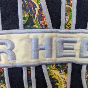 Peut inclure: Un sweat-shirt bleu marine avec un patch blanc sur lequel est brod&eacute; en bleu "HER HEART". Le patch est entour&eacute; d'un motif paisley bleu.