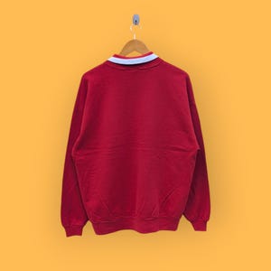 May include: Red sweatshirt with a white and red striped collar. The sweatshirt has long sleeves and a crew neck.