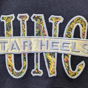 Peut inclure: Un sweat-shirt bleu marine avec un patch brod&eacute; blanc et bleu qui dit "TAR HEELS" et un motif paisley "UNC" en jaune, vert, rouge et bleu.