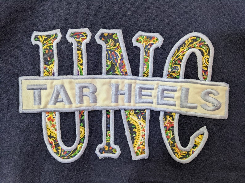 Peut inclure: Un sweat-shirt bleu marine avec un patch brod&eacute; blanc et bleu qui dit "TAR HEELS" et un motif paisley "UNC" en jaune, vert, rouge et bleu.