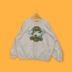 Peut inclure: Un sweat-shirt gris clair avec un grand imprimé graphique d'un poisson-chat sautant hors de l'eau. Le graphique est entouré d'un ovale doré avec le texte "Get Reel" dans une bannière en dessous.