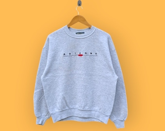 May include: A light gray sweatshirt with the word "ARIZONA" embroidered in black, with a red oval accent. The sweatshirt is hanging on a wooden hanger against a yellow background.