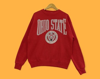 Pode incluir: Camisola de cor vermelha com um logotipo "Ohio State" branco e um emblema circular com as palavras "Ohio State University 1870" em branco.