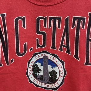 May include: Red sweatshirt with black lettering that reads "N.C. STATE" and a circular logo underneath that reads "North Carolina State University".