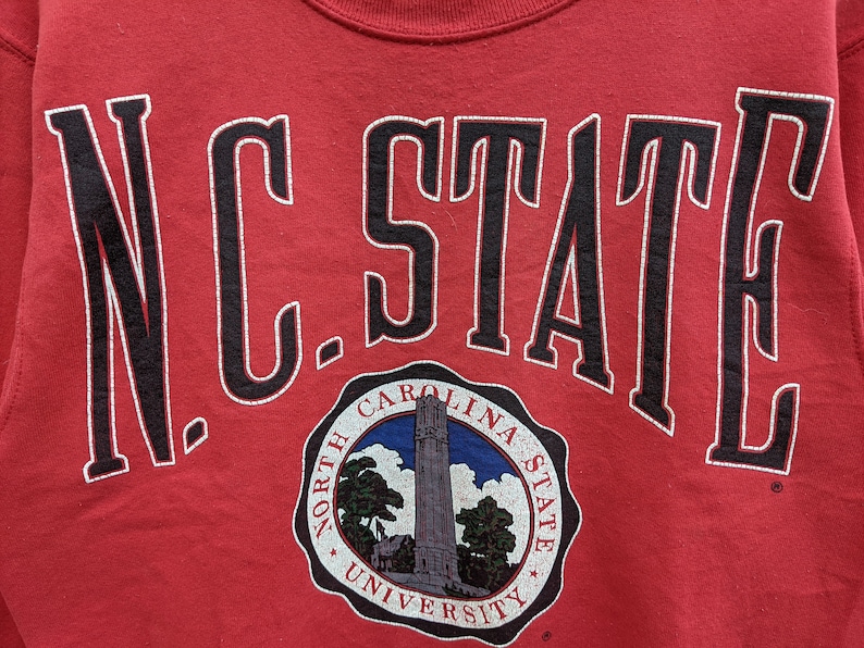 May include: Red sweatshirt with black lettering that reads "N.C. STATE" and a circular logo underneath that reads "North Carolina State University".