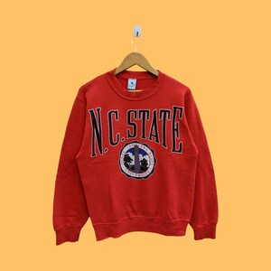 May include: Red sweatshirt with a large black and white "N.C. STATE" graphic on the front. The graphic also includes a small circular logo with the words "North Carolina State University".