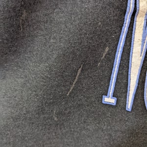 Puede incluir: Primer plano de una camiseta negra con un dise&ntilde;o gr&aacute;fico blanco y azul. La tela tiene un aspecto texturizado con algunas imperfecciones menores. El dise&ntilde;o gr&aacute;fico es parcialmente visible, con un contorno azul.