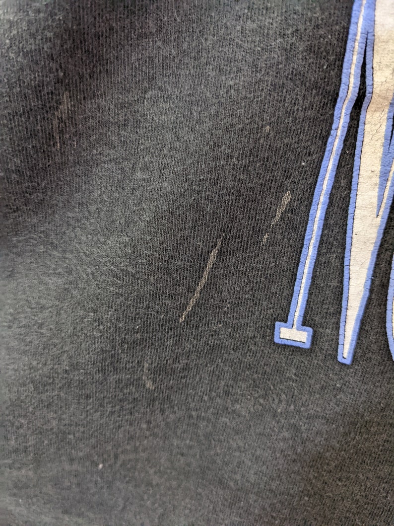 Puede incluir: Primer plano de una camiseta negra con un dise&ntilde;o gr&aacute;fico blanco y azul. La tela tiene un aspecto texturizado con algunas imperfecciones menores. El dise&ntilde;o gr&aacute;fico es parcialmente visible, con un contorno azul.