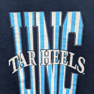 May include: A navy blue t-shirt with a white and light blue striped graphic of the word "UNC" with the words "Tar Heels" underneath.
