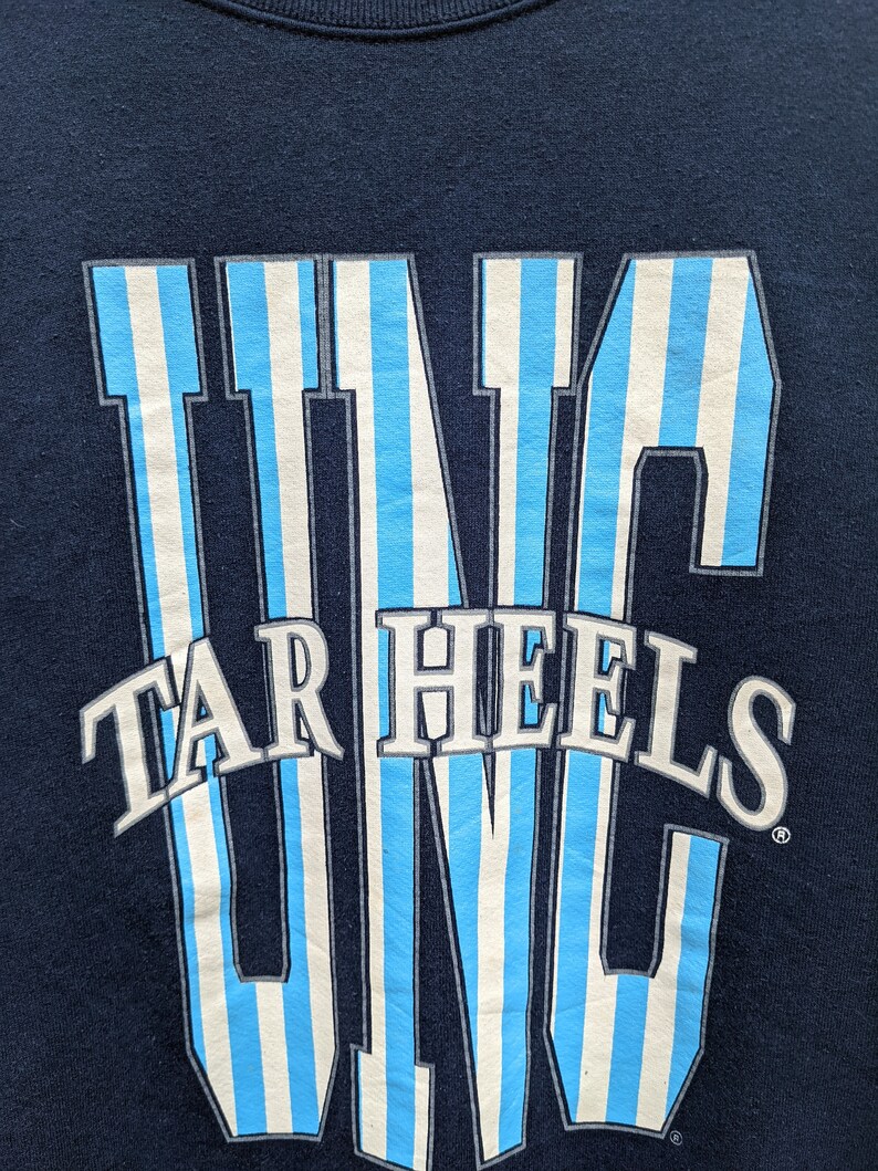 May include: A navy blue t-shirt with a white and light blue striped graphic of the word "UNC" with the words "Tar Heels" underneath.