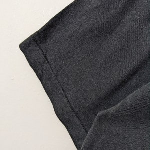 Puede incluir: Primer plano de una manga de camiseta gris oscuro. La tela tiene un aspecto texturizado, con costuras visibles a lo largo del borde. La manga est&aacute; en &aacute;ngulo, mostrando el dobladillo doblado y la textura del material.