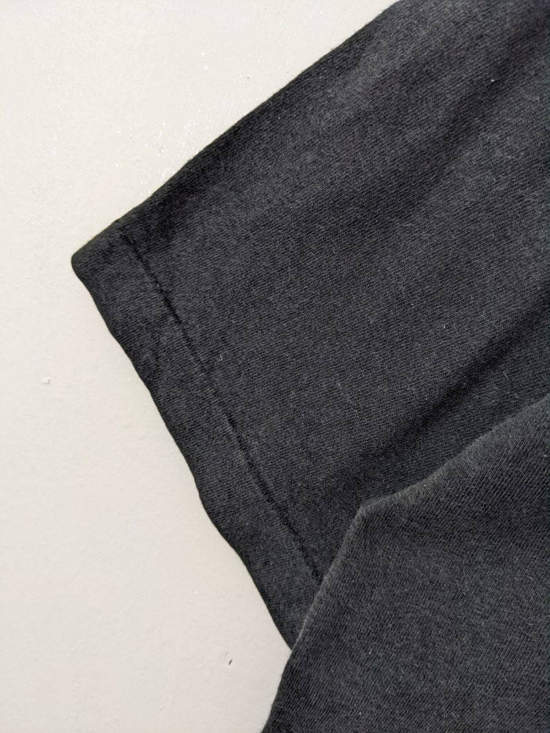 Puede incluir: Primer plano de una manga de camiseta gris oscuro. La tela tiene un aspecto texturizado, con costuras visibles a lo largo del borde. La manga est&aacute; en &aacute;ngulo, mostrando el dobladillo doblado y la textura del material.
