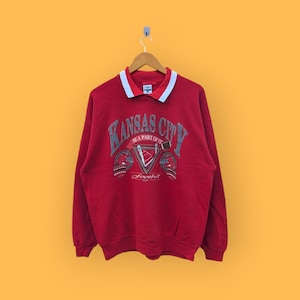 May include: Red sweatshirt with a white and blue collar. The front of the sweatshirt has a graphic print that says "Kansas City" with the words "Be a part of it" and "Football" underneath.