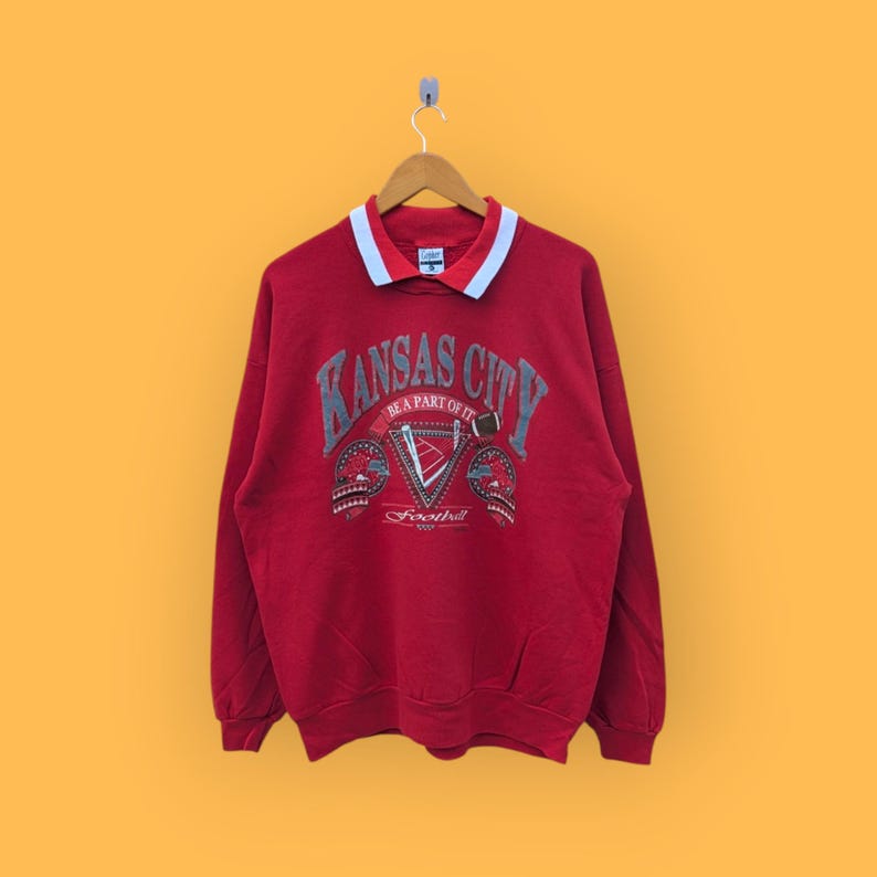 May include: Red sweatshirt with a white and blue collar. The front of the sweatshirt has a graphic print that says "Kansas City" with the words "Be a part of it" and "Football" underneath.