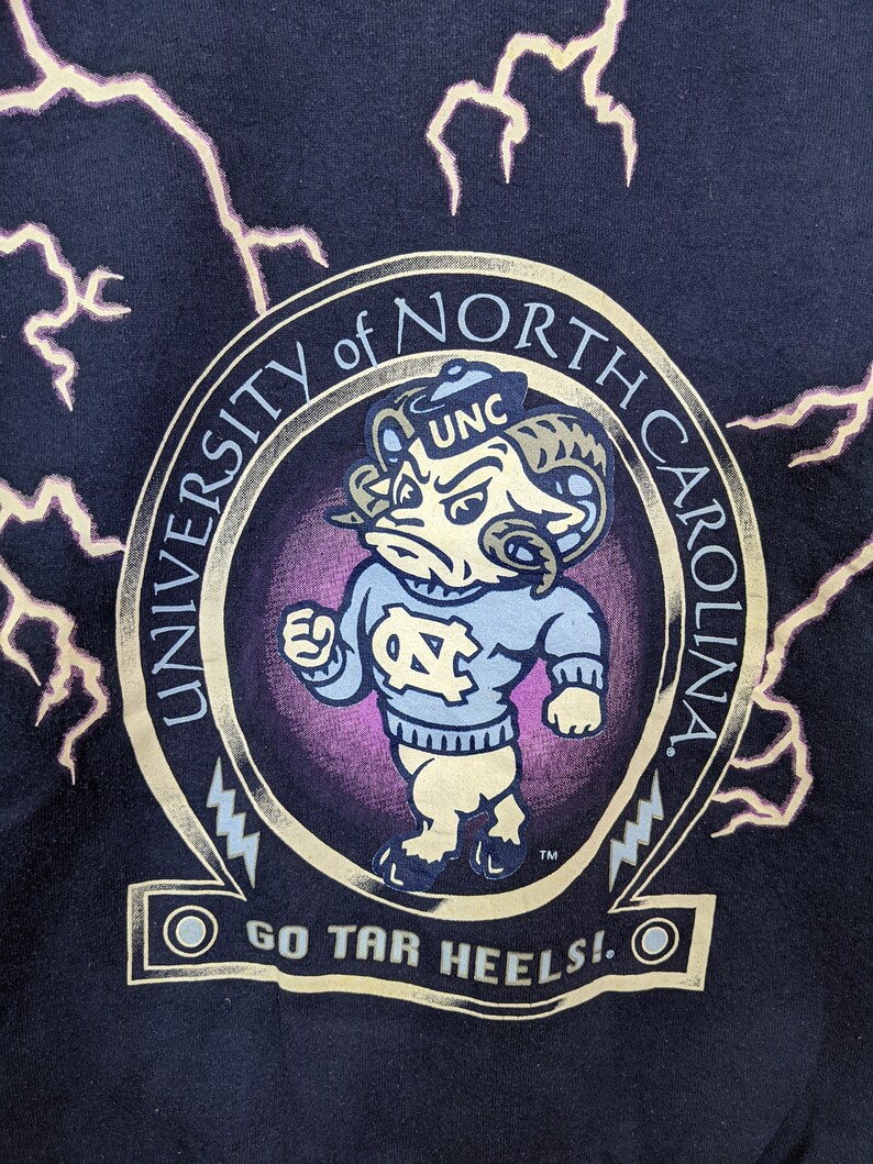 May include: Navy blue sweatshirt featuring a University of North Carolina Tar Heels graphic. The design includes a cartoon ram mascot in a light blue sweater, the words "University of North Carolina," and "Go Tar Heels!" in gold lettering, with lightning bolt accents.