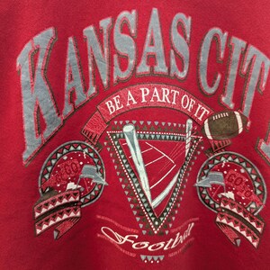 May include: Red sweatshirt with a vintage-style graphic design featuring the text "Kansas City" in a gray and white font. The design also includes a football, a stylized football field, and the text "Be a part of it" and "Football".