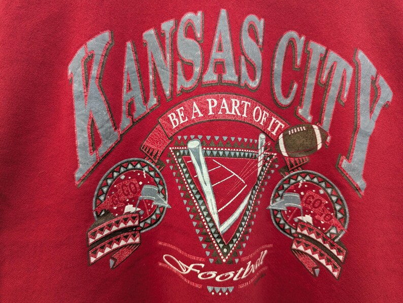 May include: Red sweatshirt with a vintage-style graphic design featuring the text "Kansas City" in a gray and white font. The design also includes a football, a stylized football field, and the text "Be a part of it" and "Football".