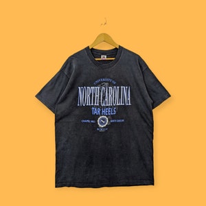 Puede incluir: Una camiseta negra con el texto "UNIVERSITY OF NORTH CAROLINA TAR HEELS" en letras blancas y azules. La camiseta tambi&eacute;n presenta un emblema circular con las palabras "CHAPEL HILL" y "NORTH CAROLINA". La camiseta est&aacute; colgada en una percha de madera.