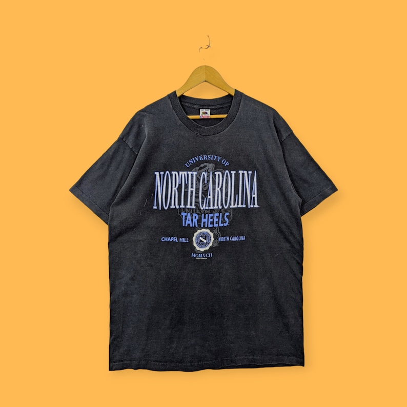 Puede incluir: Una camiseta negra con el texto "UNIVERSITY OF NORTH CAROLINA TAR HEELS" en letras blancas y azules. La camiseta tambi&eacute;n presenta un emblema circular con las palabras "CHAPEL HILL" y "NORTH CAROLINA". La camiseta est&aacute; colgada en una percha de madera.
