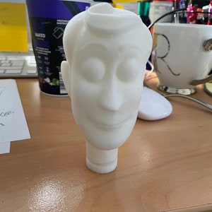 May include: White plastic 3D printed figurine of a cartoon character's head with closed eyes and a smile. The figurine is on a wooden surface.