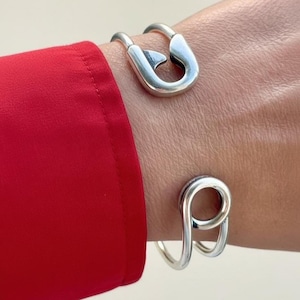 May include: Two silver-colored bracelets. The top bracelet is a safety pin design, and the bottom bracelet has a looped design. The bracelets are worn on a wrist, with a red sleeve in the background.