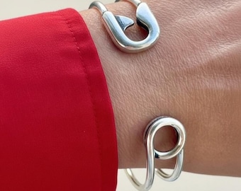 May include: Two silver bracelets. The top bracelet is a safety pin design, and the bottom bracelet has a looped design. The bracelets are worn on a wrist, with a red sleeve in the background.