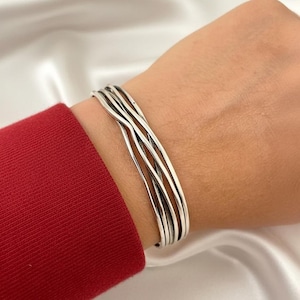 May include: A silver bracelet with a layered design. The bracelet has multiple thin, wavy bands that wrap around the wrist. The background is a soft, white fabric and a red sleeve.