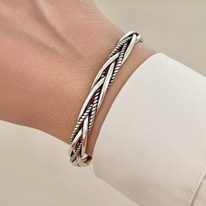 May include: A silver-colored braided bracelet. The bracelet features a rope-like design with multiple strands woven together. The bracelet is worn on a wrist, and the background includes a white shirt.