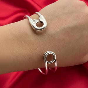 Op de afbeelding: Een zilverkleurige armband met een veiligheidsspeldontwerp. De open armband heeft een veiligheidsspeldvorm aan de bovenkant en een lus aan de onderkant. De armband zit om een pols, tegen een rode achtergrond.