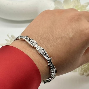 May include: A silver-toned bracelet featuring a wing design. The bracelet is worn on a wrist, with the wings curving around the wrist. The wings have detailed feather patterns. The background includes a red sleeve and white flowers.