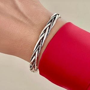 May include: A silver-colored braided cuff bracelet. The bracelet features a woven design with multiple strands, creating a textured appearance. The bracelet is worn on a wrist, with a red garment in the background.