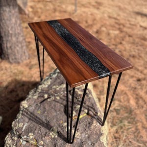 May include: A walnut wood table with a black epoxy river inlay and hairpin legs. The table is sitting on a large rock.