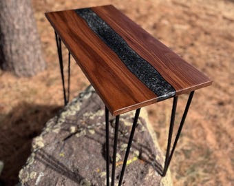 May include: A walnut wood table with a black epoxy river inlay and hairpin legs. The table is sitting on a large rock.