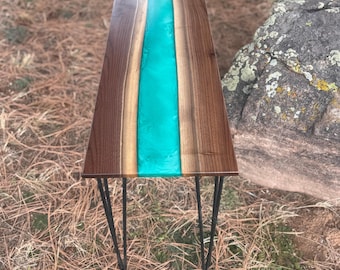 May include: A rectangular wooden table with a turquoise resin river down the center. The table has dark brown wood on either side of the resin and black hairpin legs. The table is approximately 36 inches long.