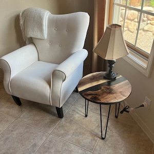 May include: A white armchair with a throw blanket sits next to a small, round, wooden side table with black metal legs. A lamp with a beige lampshade sits on the table.