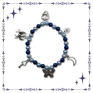 Puede incluir: Una pulsera de cuentas con abalorios plateados. La pulsera presenta cuentas azul claro y azul oscuro alternadas. Los abalorios incluyen una calavera, una araña, una llave, una mariposa, una luna creciente y un diseño en forma de corazón.