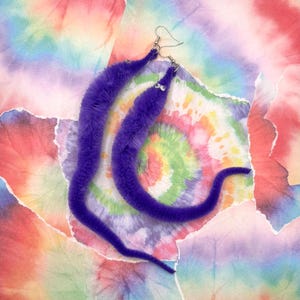May include: A pair of quirky purple worm earrings with silver ear wires. The earrings are made of a fuzzy material and have small decorative accents. The background features a colourful tie-dye pattern.