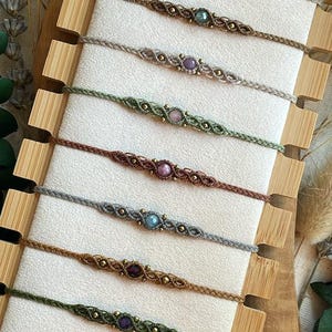 May include: A collection of twelve thin macrame bracelets arranged vertically on a light-coloured display board. Each bracelet features a central round gemstone bead flanked by small metallic accents, crafted from various coloured cords.