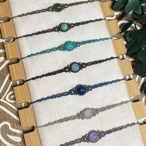 May include: Ten macrame bracelets arranged in a vertical row on a display board. Each bracelet features a single round gemstone in various colors including red, peach, green, turquoise, blue, lavender, and gray, accented with small metallic beads.