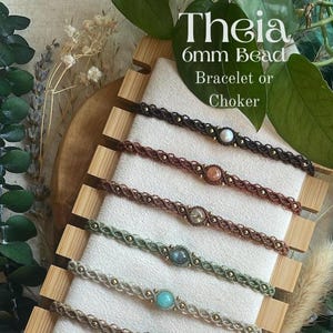 May include: A display of handmade macrame bracelets or chokers with 6mm beads. The bracelets are in various colors, including brown, green, and blue, each featuring a central gemstone. The text "Theia" and "Bracelet or Choker" are visible.