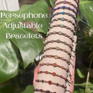 May include: A collection of Persephone Adjustable Bracelets featuring brown cord bands with single faceted beads in various colours, including pink, blue, green, orange, and red, arranged vertically on a beige cylindrical display stand.
