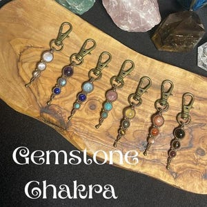 May include: Seven metal lobster clasp charms featuring various round gemstones arranged in vertical patterns. The text Gemstone Chakra Charms is printed in white. Each charm is constructed with wire-wrapped beads and a bronze-toned metal clip.