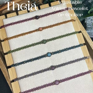 May include: Six adjustable anklet, bracelet, or choker pieces in various colors including dark purple, orange, olive green, teal, grey, and light purple. Each features a central round gemstone and small metallic beads on a woven cord.