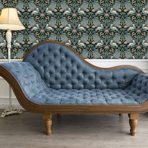 May include: A blue tufted chaise lounge with wooden legs and a wooden frame. The chaise lounge is in front of a wall with a floral patterned wallpaper.