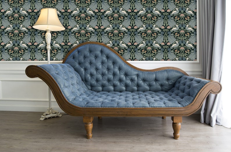 May include: A blue tufted chaise lounge with wooden legs and a wooden frame. The chaise lounge is in front of a wall with a floral patterned wallpaper.