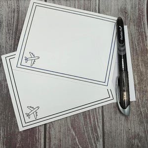 May include: Two white notecards with a blue airplane graphic and a black border are stacked on a wooden surface. A black and silver pen rests on the notecards. The pen has the words "Fountain Pen" printed on it.