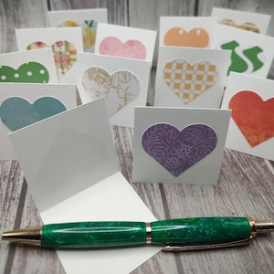 May include: A collection of handmade greeting cards with heart-shaped cutouts in various colors and patterns. A green and gold pen rests in front of the cards. The cards are displayed against a light wood background.