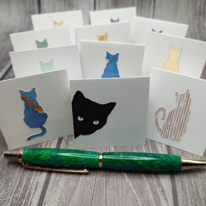 May include: A green and gold pen rests in front of several white cards with cat silhouettes in various colors. The pen has a marbled green barrel and gold accents. The cards feature blue, yellow, and black cat designs.