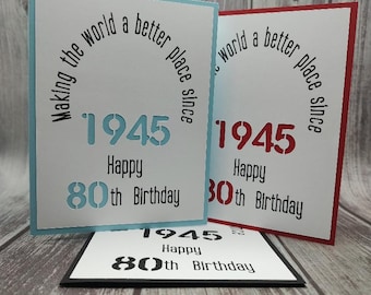 May include: Three rectangular greeting cards featuring the text "Making the world a better place since 1945" and "Happy 80th Birthday" in a stencil font. One card has a light blue border, one has a red border, and one has a black border.